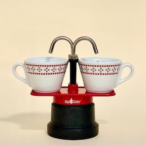 Könnte beinhalten: Ein roter und schwarzer Espressokocher mit zwei weißen Tassen. Die Tassen haben ein rot-schwarzes Muster. Der Espressokocher hat einen silbernen Ausguss und ist zum Brühen von Kaffee konzipiert.