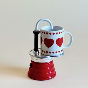 May include: A red and silver stovetop espresso maker with a white mug featuring red hearts. The mug sits atop the espresso maker, which has a curved silver spout and a black handle. The design is simple and charming.