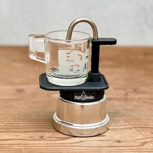 May include: A small, silver espresso maker with a clear glass cup. The cup has a handle and text on the side. A curved metal spout extends from the top. The espresso maker sits on a wooden surface.
