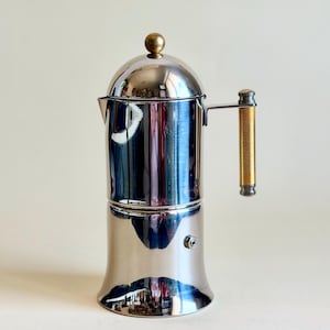 Puede incluir: Una cafetera de acero inoxidable pulido con cuerpo cilíndrico, un pequeño pico vertedor y una tapa abovedada rematada con un pomo esférico de tono dorado. El lateral presenta un asa vertical con un agarre texturizado de tono dorado y detalles metálicos.