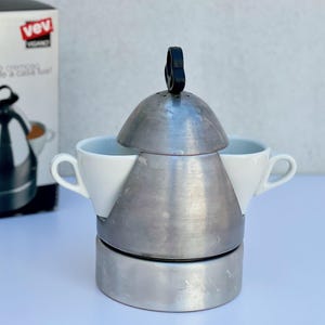 Może przedstawiać: Biały ceramiczny kubek z czarnym uchwytem i napisem "But First, Coffee" pogrubioną czcionką. Kubek jest ustawiony na rozmytym tle, sugerującym przytulne otoczenie.