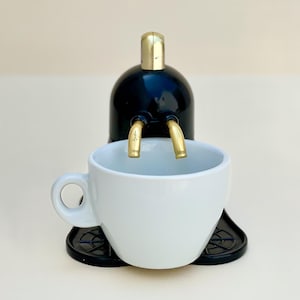 Op de afbeelding: Een kleine espressomachine met een zwarte behuizing, gouden accenten en een witte keramische kop. De kop staat onder de tuit. Het ontwerp is modern en compact. Geschikt voor een enkele portie.