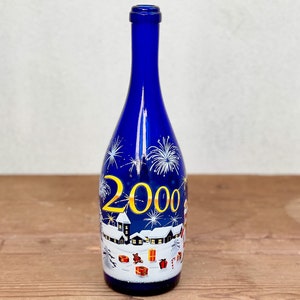 Puede incluir: Una botella de vidrio azul cobalto con una escena invernal pintada a mano que muestra casas cubiertas de nieve, cajas de regalo dispersas y fuegos artificiales blancos. El número 2000 aparece en grandes cifras amarillas en el centro.