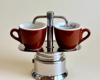 Op de afbeelding: Een zilverkleurige metalen espressomaker-standaard met twee kleine bruine keramische kopjes met een witte binnenkant. Het rechterkopje bevat de tekst "Battista Caffe" in cursief schrift. De standaard heeft een centrale paal met twee tuiten.