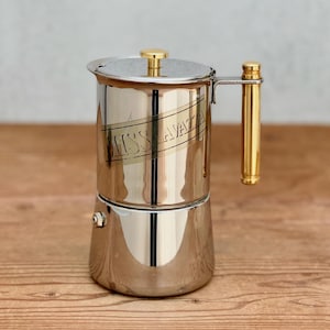 Peut inclure: Une cafetière italienne en acier inoxydable poli dotée d'une poignée cylindrique dorée et d'un bouton assorti sur le couvercle. Le côté de la chambre supérieure porte l'inscription gravée MISS LAVAZZA disposée en diagonale.
