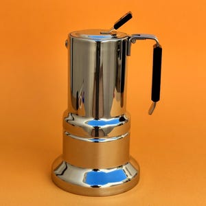 Può includere: Una caffettiera in acciaio inox lucido, con manico e leva del coperchio neri. La caffettiera è cilindrica con base arrotondata, su sfondo arancione. Un classico elettrodomestico da cucina.