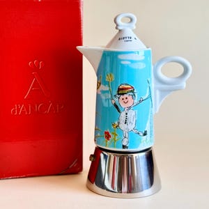 以下が含まれることがあります： 帽子をかぶった人物のカラフルなイラストが描かれた白と水色のセラミック製モカポットと、A d'ANCAPのエンボス文字が入った赤い長方形の箱。ポットには金属製のベース、白いハンドル、白い蓋が付いています。