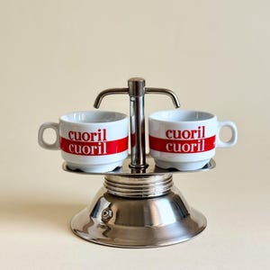 Può includere: Una caffettiera in acciaio inossidabile con due tazze bianche. Ogni tazza ha una striscia rossa con la scritta "cuoril". La caffettiera è su uno sfondo beige chiaro.