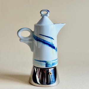 May include: A white ceramic stovetop espresso maker with a stainless steel base. The white body features blue and yellow brushstroke designs. The handle and lid knob have blue accents. A classic coffee brewing appliance.