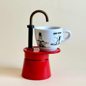 Könnte beinhalten: Ein leuchtend roter Espressokocher-Sockel mit einem schwarzen vertikalen Pfosten und einem gebogenen messingfarbenen Auslauf. Eine weiße Keramik-Espressotasse mit einer schwarzen Grafik einer Person und einer Kaffeekanne mit dem Text Moka Express.