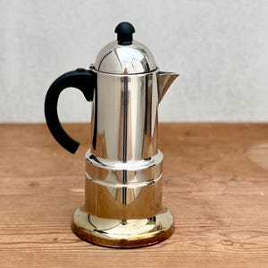 May include: A shiny, stainless steel stovetop espresso maker with a black handle and a black knob on top. The coffee pot is sitting on a wooden surface.