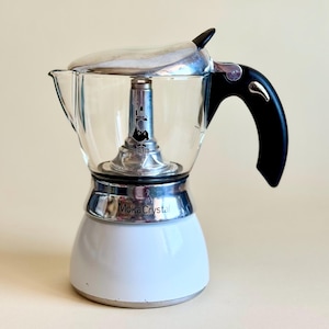 Puede incluir: Una cafetera italiana Moka Crystal con cámara superior de cristal transparente, base blanca y asa negra. La columna central de metal muestra el logotipo de Bialetti y el texto Moka Crystal está impreso en la banda plateada.
