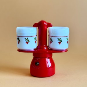 Peut inclure: Une cafetière rouge avec deux tasses blanches. Chaque tasse présente un motif de cerise rouge et de feuilles vertes. La cafetière a une base ronde et une poignée sur le dessus. Le fond est de couleur beige.