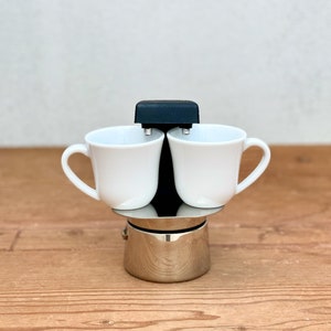 Peut inclure: Une base de cafetière italienne en acier inoxydable avec une colonne centrale noire et deux petites tasses en céramique blanche placées de chaque côté. L'appareil présente une finition métallique polie et un élément supérieur en plastique noir.