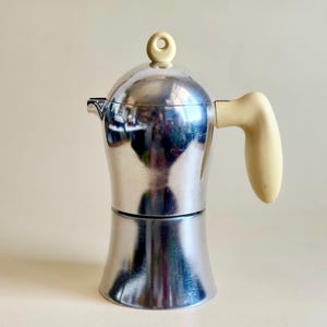 Könnte beinhalten: Ein polierter, silberfarbener Espressokocher aus Metall mit einem glatten, abgerundeten Körper. Er verfügt über einen cremefarbenen, ergonomischen Griff und einen passenden cremefarbenen, runden Knopf auf dem Deckel. Die Oberfläche ist reflektierend.