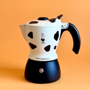 Puede incluir: Una cafetera espresso en blanco y negro con un diseño de vaca. La cafetera tiene un asa negra y una base negra. El texto "Mukka Express" está impreso en la parte delantera.