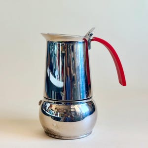 May include: A polished stainless steel stovetop espresso maker with a rounded base and a cylindrical upper chamber. The coffee pot features a bright red curved handle attached to the side and a small metal lid hinge at the top rim.