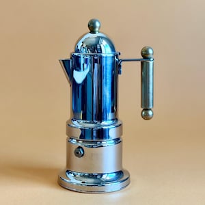 May include: A shiny, chrome-finished stovetop espresso maker. The coffee pot has a round base, a cylindrical body, and a lid with a small knob. It features a handle and a spout for pouring. The espresso maker is a classic kitchen appliance.