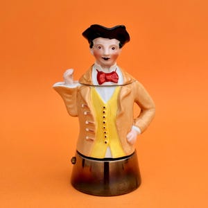 Peut inclure: Figurine en céramique représentant un homme en veste beige, gilet jaune et nœud papillon rouge. La figurine a un chapeau noir et une base marron. Le fond est de couleur orange.
