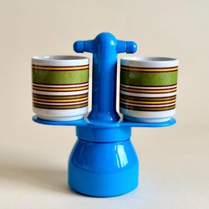 May include: A bright blue coffee maker with a central handle and two matching cups resting on side platforms. Each cup features horizontal stripes in shades of green, brown, yellow, and white. The coffee maker has a smooth, glossy finish.