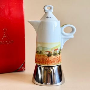 Op de afbeelding: Een witte en zilveren espressomaker met een landschapsontwerp. De espressomaker heeft een handvat en een deksel. Een rode doos met de merknaam "D'ANCAP" staat links.