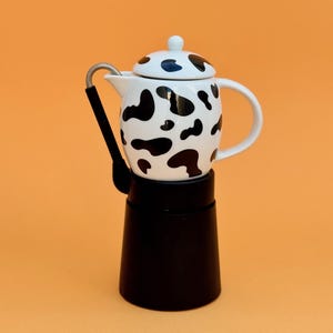 Può includere: Una caffettiera espresso in bianco e nero con un motivo a mucca. La parte superiore è bianca con macchie nere e la base è nera. Il manico è nero e argento. Lo sfondo è arancione.