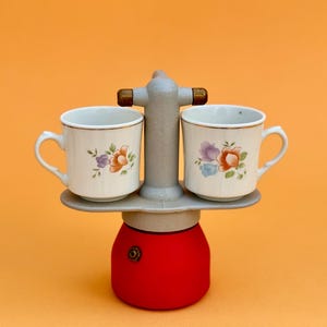 Può includere: Una caffettiera rossa e grigia con due tazze decorate con fiori. Le tazze sono color crema con un manico e un motivo floreale arancione, viola e verde. La caffettiera ha una base rossa e una parte superiore grigia.