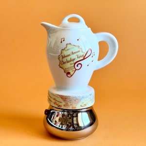 Puede incluir: Una cafetera de cerámica blanca con asa y tapa, que presenta una etiqueta decorativa con el texto Johann Strauss Der lustige Krieg y notas musicales. La base es un componente metálico plateado pulido con un acabado reflectante.