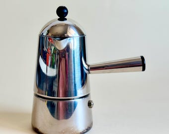 May include: A polished stainless steel stovetop espresso maker with a cylindrical body, a rounded lid featuring a small black knob, and a side-mounted handle. The coffee pot has a tapered shape, a small pouring spout, and a metallic finish.