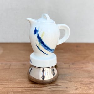 Pode incluir: Uma cafeteira italiana com base em aço inoxidável polido e parte superior em cerâmica branca. A secção de cerâmica inclui uma pega, uma tampa com um pequeno botão e um design abstrato pintado em tons de azul e amarelo.