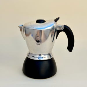 Puede incluir: Una cafetera Moka Express plateada y negra con asa y tapa negras. La cafetera tiene el logotipo de Bialetti y las palabras "Mukka Express" impresas. La cafetera está diseñada para preparar café estilo espresso.