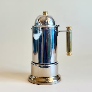 Pode incluir: Uma cafeteira de cromo polido com corpo cilíndrico, tampa arredondada com um pequeno botão dourado e uma alça vertical com detalhes dourados na parte superior e inferior. A superfície metálica reflete a luz com muita clareza.