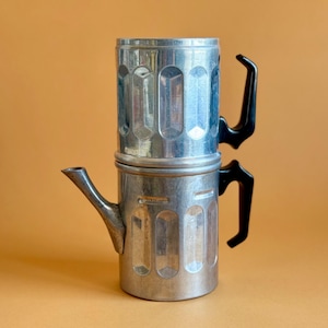 Pode incluir: Uma cafeteira prateada vintage com uma alça e um bico pretos. A cafeteira é feita de metal e tem um design de duas partes. O fundo é de cor laranja lisa.