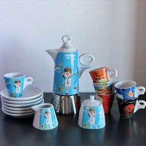 Op de afbeelding: Een grillige koffieset met een blauw en wit ontwerp met een stripfiguur. De set bevat een espressomaker, kopjes, schotels, een melkkan en een suikerpot. De kopjes hebben diverse kleurrijke ontwerpen. Afmetingen in centimeters.