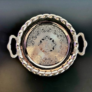 May include: A round silver-toned metal serving tray featuring two ornate handles on opposite sides. The tray has a scalloped edge and a central circular area decorated with intricate, symmetrical floral and scrollwork engravings.