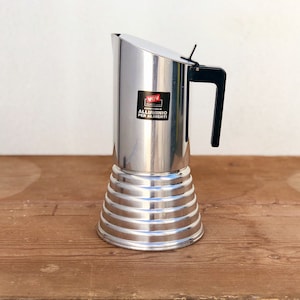May include: A stainless steel stovetop espresso maker with a black handle and a ridged base. The coffee pot has a label with Italian text. The pot is sitting on a wooden surface.