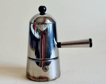 May include: A polished stainless steel stovetop espresso maker with a cylindrical body, a rounded lid featuring a small black spherical knob, and a side handle with a black end cap. A small metal pressure valve is visible on the lower base.
