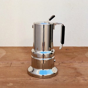Op de afbeelding: Een gepolijste roestvrijstalen espressomaker met een zwarte handgreep en knop. De koffiepot heeft een reflecterend oppervlak en staat op een houten oppervlak.
