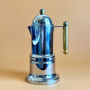May include: A shiny, chrome-plated stovetop espresso maker with a gold-colored handle and accents. The coffee pot has a rounded top and base, with a spout for pouring. The handle is cylindrical and attached to the side of the pot.