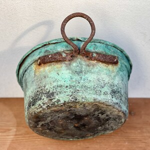 Può includere: Un contenitore metallico rotondo e usurato, caratterizzato da una finitura con patina verde acqua. Il recipiente presenta un manico circolare in ferro arrugginito, fissato al bordo superiore con una staffa metallica rettangolare.