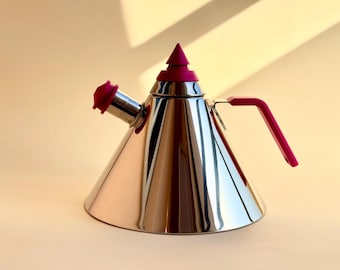 Op de afbeelding: Een roestvrijstalen waterkoker met een conische body en een magenta handvat, tuitdop en bovenkant. De waterkoker heeft een gepolijst, reflecterend oppervlak en het handvat is een eenvoudig, hoekig ontwerp. De algehele vorm is modern en strak.