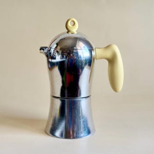 Pode incluir: Uma cafeteira de metal prateado brilhante para fogão com uma alça arredondada na cor creme e um botão correspondente na tampa. A cafeteira tem uma base larga que se estreita para cima com um bico e uma tampa em formato de cúpula.
