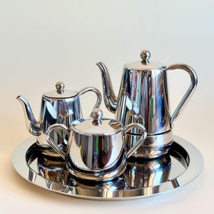May include: A polished silver-tone metal tea set consisting of a tall teapot, a smaller teapot, and a lidded sugar bowl with two handles. All three pieces are arranged on a matching round, reflective silver-tone serving tray with a smooth rim.