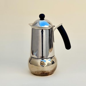 May include: A polished stainless steel stovetop espresso maker with a rounded base, a cylindrical upper chamber, and a black heat-resistant handle. The lid features a small black knob, and a brass-colored pressure valve is visible on the side.