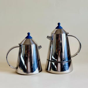 Op de afbeelding: Twee roestvrijstalen espressopotten met blauwe knoppen. De koffiepotten hebben een klassiek ontwerp met een handvat en tuit. Het glanzende metaal reflecteert de omgeving. De ene is kleiner dan de andere.