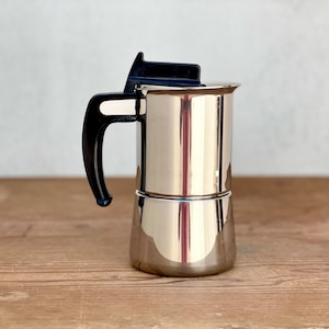 Op de afbeelding: Een roestvrijstalen espressomaker met een zwarte handgreep en deksel. De koffiepot is cilindrisch met een tweedelig ontwerp en staat op een houten oppervlak. De handgreep is gebogen en aan de zijkant bevestigd.