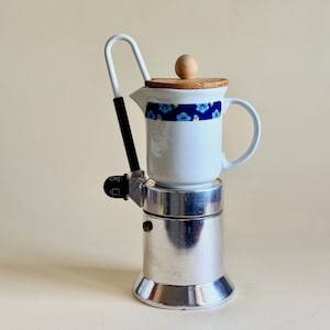 Puede incluir: Una cafetera blanca con estampado floral azul, tapa de madera y base plateada. El asa es blanca con agarre negro. La cafetera está diseñada para uso en estufa.