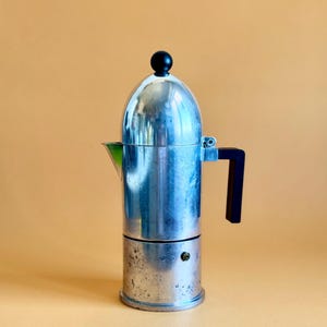 Può includere: Una caffettiera argentata da piano cottura con manico nero e pomello nero sulla parte superiore. La caffettiera ha un beccuccio ed è in metallo. La caffettiera è su uno sfondo arancione chiaro.