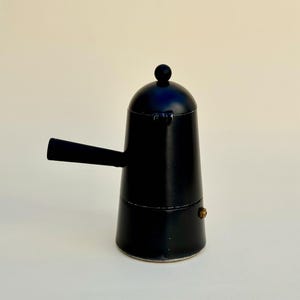 May include: A black stovetop espresso maker with a rounded top, a handle, and a small knob. The coffee maker is made of metal and has a classic design. The espresso maker is approximately 7 inches tall.