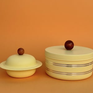 May include: Two pale yellow ceramic containers with wooden knobs, set against an orange backdrop. One is a small, lidded dish. The other is a larger, round container with silver bands. These are likely storage containers.